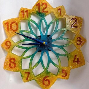 May include: A wall clock with a star-shaped frame featuring a gradient of yellow, orange, and teal colors with embedded glitter. The clock face has cut-out petal patterns, raised orange numbers from one to twelve, and two dark blue clock hands.