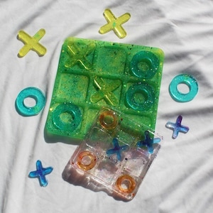 May include: Two green and clear resin tic-tac-toe game boards with colorful X and O pieces. The green board has glitter embedded in the resin. The clear board has a pearlescent finish.