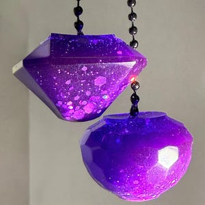 May include: Two purple resin ornaments with faceted surfaces and embedded glitter particles. One ornament is diamond-shaped, and the other is spherical. Both items are suspended from dark metal ball chains, creating a reflective and translucent appearance.
