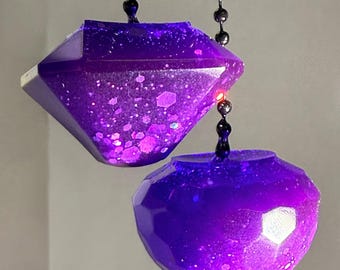 Puede incluir: Dos adornos de resina morada con superficies facetadas y partículas de purpurina incrustadas. Un adorno tiene forma de diamante y el otro es esférico. Ambos artículos están suspendidos de cadenas de bolas de metal oscuro, creando un efecto.