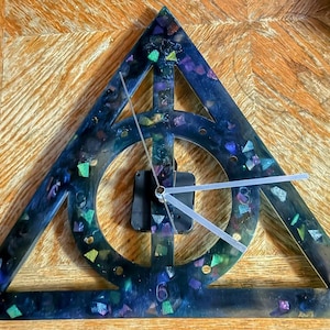 May include: A black triangle clock with a white clock face and silver hands. The triangle is filled with iridescent glitter and has a circle in the center. The clock is on a wooden surface.