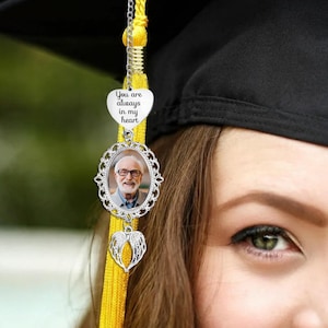 May include: A silver graduation tassel charm with a photo of a person in glasses, a heart-shaped pendant with the text "You are always in my heart", and a small pair of angel wings. The charm hangs from a yellow tassel.