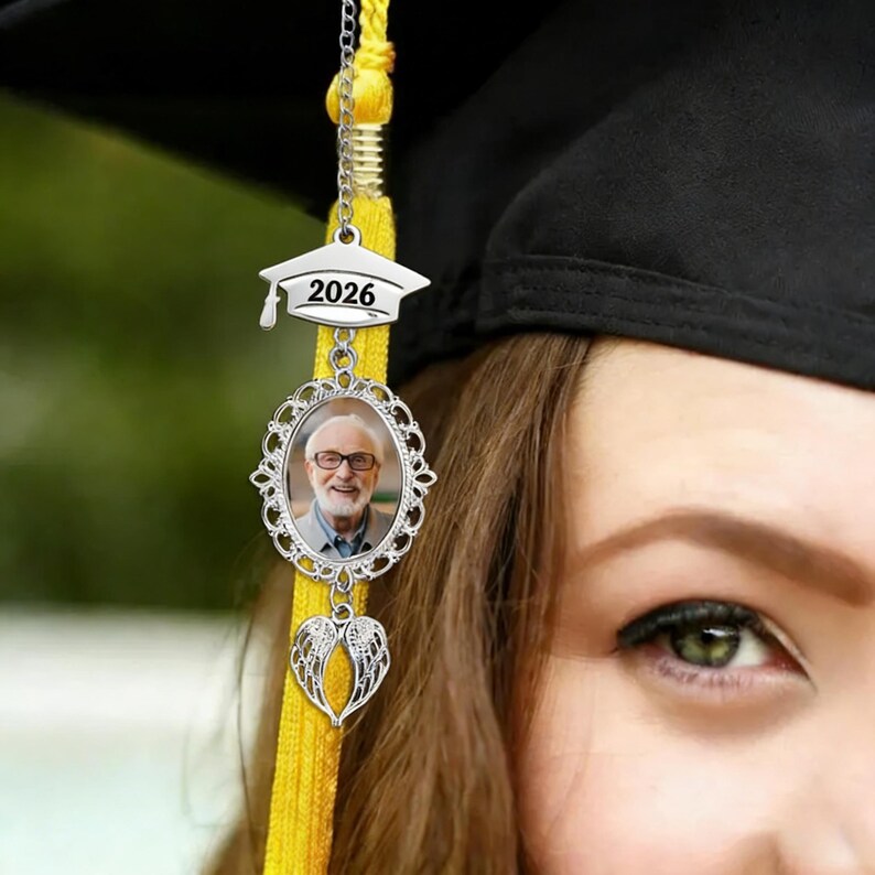 May include: A graduation cap tassel charm with a silver-colored graduation cap pendant displaying the year 2026. Below is an oval frame with a photo of a person, and a heart-shaped charm. The tassel is yellow.