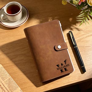 May include: A brown leather journal with a snap closure, the name "WILLIAMS" is printed in black. A black and gold pen rests beside the journal. A cup of tea sits on a saucer.