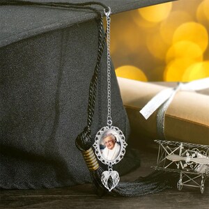 May include: A graduation cap with a black tassel and a silver charm. The charm features a framed photo of a person, a decorative border, and a heart with angel wings. The background is blurred with a graduation scroll.