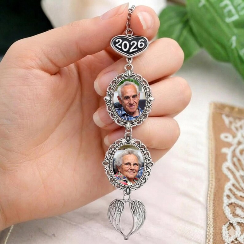 May include: A silver-colored memorial pendant with two oval photo frames and a heart-shaped top with the year 2026. The bottom of the pendant features a pair of angel wings.