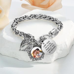 Może przedstawiać: Srebrna bransoletka z plecionym wzorem łańcucha. Bransoletka posiada okrągły charms ze zdjęciem i napisem "I Will Hold You In My Heart UNTIL I HOLD YOU IN HEAVEN". Zawiera skrzydła i ozdobne zawieszki.