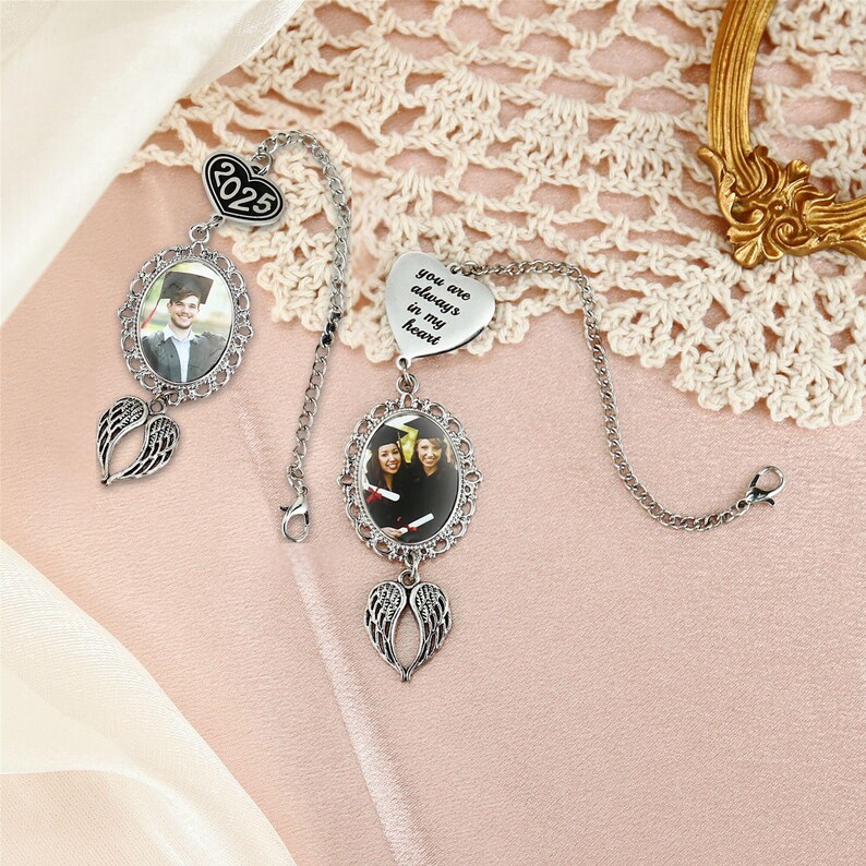 May include: Two graduation-themed car charms. One features a photo in an oval frame with angel wings, a chain, and a charm with the year 2025. The other has a heart charm with the text "you are always in my heart" and a photo in an oval frame with angel wings.
