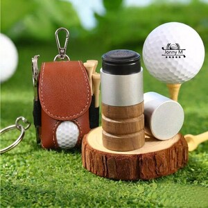 May include: A brown leather golf ball holder with a silver carabiner clip, a golf ball, and a golf ball marker. The golf ball has the text "Jonny M" on it. The holder also contains golf tees and a divot repair tool.