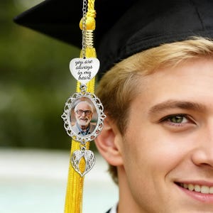 May include: A silver graduation tassel charm with a photo of a man in glasses, a heart-shaped pendant with the words "you are always in my heart", and a heart with angel wings. The tassel is yellow.