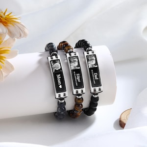May include: Three beaded bracelets with rectangular silver pendants featuring black and white photos. Each pendant is engraved with a name: "Mom", "Edgar", and "Dad". The bracelets have different colored beads: black, brown, and gray.