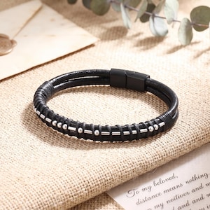 May include: A black leather bracelet featuring a double-strand design with a matte black magnetic clasp. The band is accented with a series of silver-toned metal beads and short bars arranged in a pattern along the leather cord surface.