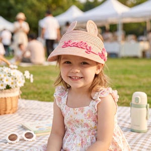 May include: A light pink straw sun visor featuring two upright ears on the crown, a pink embroidered script text that reads Sally, and a small pink flower accent on the side. The visor has a wide brim and a woven texture throughout the material.
