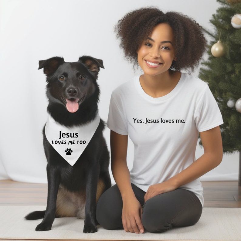 Puede incluir: En esta imagen aparecen una mujer y un perro negro. La mujer lleva una camiseta blanca con el texto "Yes, Jesus loves me". El perro lleva un pa&ntilde;uelo blanco con el texto "Jesus loves me too" y una huella de pata.