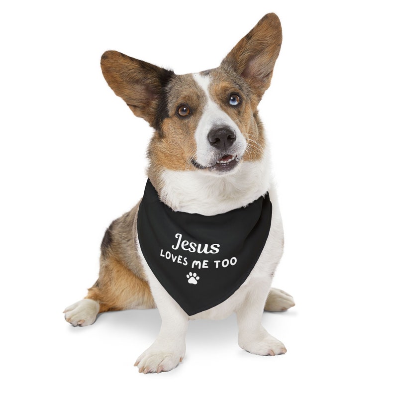 Puede incluir: Un perro Corgi tricolor con un pa&ntilde;uelo negro con el texto "Jesus LOVES ME TOO" y una huella de pata. El perro tiene un ojo azul y otro marr&oacute;n, sobre un fondo blanco.