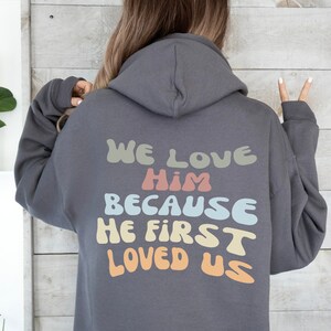 May include: A charcoal gray hoodie with a hood and long sleeves. The back of the hoodie features the text "WE LOVE HIM BECAUSE HE FIRST LOVED US" in pastel colors. The hoodie is made of a soft material.