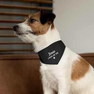 Puede incluir: Un perro Jack Russell Terrier con un pa&ntilde;uelo negro. El pa&ntilde;uelo tiene texto blanco que dice "Jesus LOVES ME TOO" con una huella de pata. El perro tiene pelo blanco, marr&oacute;n y negro. El perro est&aacute; sentado en una superficie marr&oacute;n.