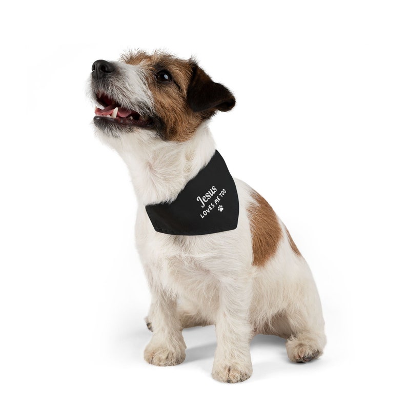 Puede incluir: Un perro peque&ntilde;o con un pa&ntilde;uelo negro con el texto "Jesus loves me too" y una huella de pata. El perro tiene pelo blanco, marr&oacute;n y negro y est&aacute; mirando hacia arriba con la boca abierta.