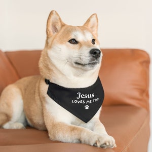 Puede incluir: Un perro Shiba Inu con un pa&ntilde;uelo negro con el texto "Jesus Loves Me Too" en blanco. El pa&ntilde;uelo tambi&eacute;n presenta una huella de pata blanca. El perro est&aacute; acostado en un sof&aacute; de cuero marr&oacute;n.