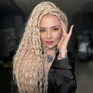 May include: A person with long, blonde, curly hair and intricate braids. They are wearing a black satin top and have vibrant makeup with yellow and pink eyeshadow. Tattoos are visible on their arm.