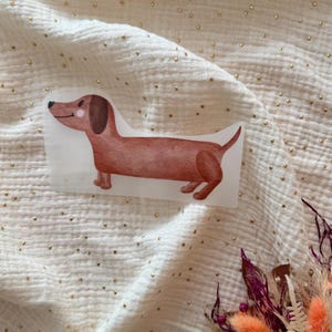 Könnte beinhalten: Ein Aufkleber im Aquarellstil eines Dackelhundes in Brauntönen. Der Hund ist im Profil dargestellt, mit einem langen Körper und kurzen Beinen. Der Aufkleber befindet sich auf einem weißen Hintergrund mit einem strukturierten Stoff.