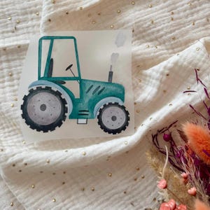 Könnte beinhalten: Ein türkisfarbener Traktoraufkleber mit grauen Rädern und einem schwarzen Schornstein. Der Traktor hat einen Fahrersitz und ein Lenkrad. Der Aufkleber befindet sich auf einem weißen Hintergrund mit Goldflocken und Trockenblumen.