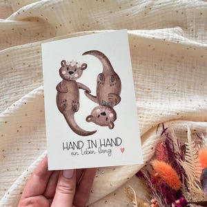 May include: A greeting card featuring watercolor illustrations of three otters. The otters are brown with floral accents. The card has the text "HAND IN HAND ein Leben lang" and a small pink heart.
