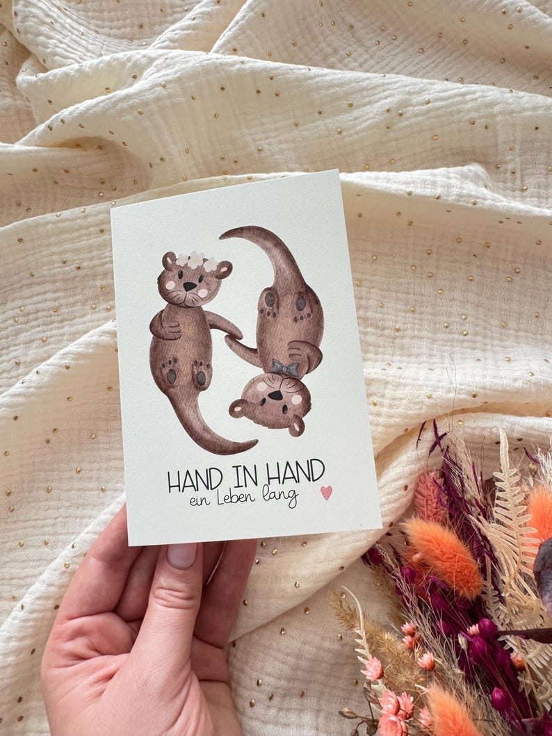 May include: A greeting card featuring watercolor illustrations of three otters. The otters are brown with floral accents. The card has the text "HAND IN HAND ein Leben lang" and a small pink heart.
