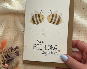May include: A greeting card with two cartoon bees, yellow and brown striped, facing each other. The card reads "You BEE-LONG together" in black script. The card is on a brown envelope.