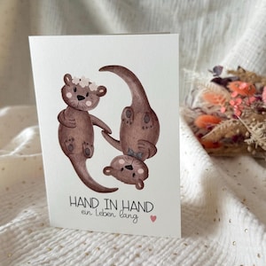 May include: A greeting card featuring three brown otter illustrations. One otter stands upright, another is upside down, and the third is between them. The card has the text "HAND IN HAND ein Leben lang" with a small pink heart.