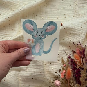 Könnte beinhalten: Aquarellillustration einer blauen Maus, die einen Blumenstrauß hält. Die Maus hat große Ohren und einen langen Schwanz. Der Hintergrund ist ein cremefarbener Stoff mit goldenen Flecken.