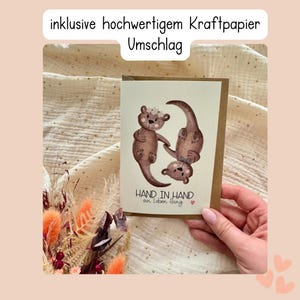 May include: A greeting card with a watercolor illustration of three brown otters. The otters are arranged in a playful pose, with one standing upright, one upside down, and one in between. The card has the text "HAND IN HAND" and "an leben lang".