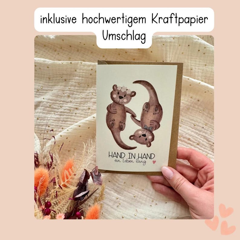 May include: A greeting card with a watercolor illustration of three brown otters. The otters are arranged in a playful pose, with one standing upright, one upside down, and one in between. The card has the text "HAND IN HAND" and "an leben lang".