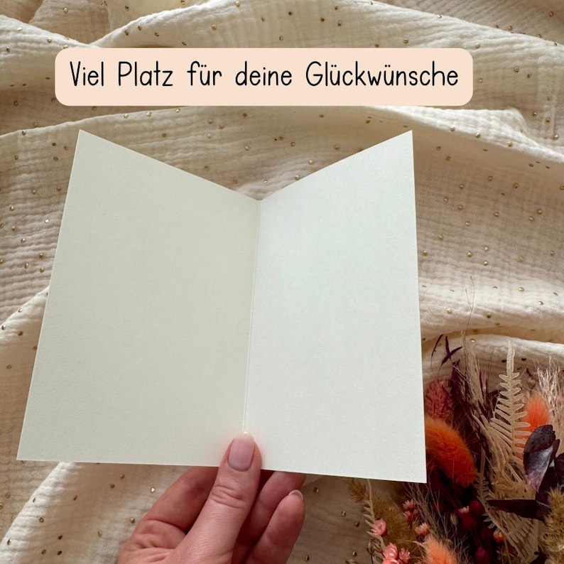 May include: An open, blank greeting card with the text "Viel Platz f&uuml;r deine Gl&uuml;ckw&uuml;nsche" in a light peach banner. The card is held open, revealing the blank interior. The background is a textured, light-colored fabric.