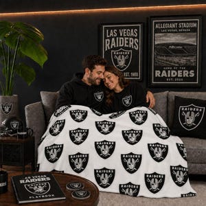 Puede incluir: Una manta de forro polar blanca con un patrón repetitivo del logotipo de los Las Vegas Raiders en negro y plata. El emblema en forma de escudo muestra a un pirata con un parche en el ojo y espadas cruzadas, con la palabra RAIDERS arriba.