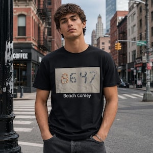 May include: Black t-shirt with a rectangular graphic featuring the numbers "8647". The number "86" is made of small, tan objects, and "47" is in dark blue. The text "Beach Comey" is below the graphic.