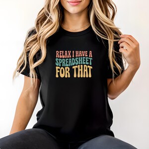 May include: A black short-sleeved t-shirt featuring the text "RELAX I HAVE A SPREADSHEET FOR THAT" printed in a stacked, wavy font. The word "RELAX" is in a muted pink, "I HAVE A" is in a muted teal, "SPREADSHEET" is in teal, and "FOR THAT" is in yellow.