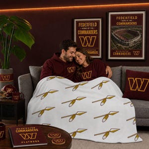May include: A white fleece blanket featuring a repeating pattern of gold and burgundy spear icons. The blanket is draped over a gray sofa, accompanied by a burgundy throw pillow with a gold W logo and a notebook labeled Washington Commanders Playbook.