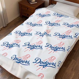 Może przedstawiać: Biały koc z powtarzającym się wzorem niebieskiego logo Dodgers oraz czerwonymi grafikami piłek baseballowych z liniami ruchu. Wzór jest nadrukowany na całej powierzchni tkaniny, która jest rozłożona na ramie łóżka.
