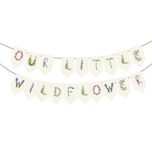 May include: A banner with the words "OUR LITTLE WILDFLOWER" spelled out on individual cream-colored pennants. Each letter is decorated with floral designs in various colors, including pink, purple, and green. The banner is strung on a thin green string.