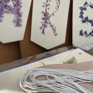 May include: A banner with the letters "A", "R", and "S" formed by purple and blue flowers. The banner is made of white paper and strung with white twine. The banner is on a wooden surface.