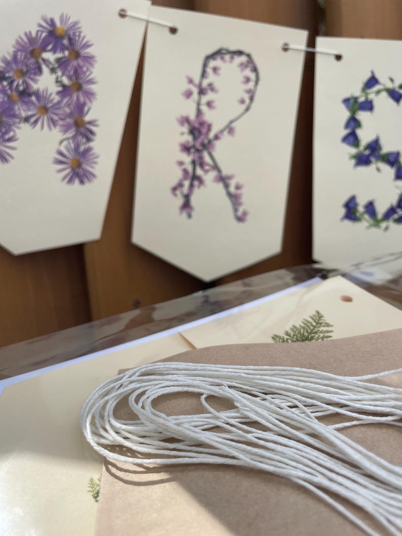 May include: A banner with the letters "A", "R", and "S" formed by purple and blue flowers. The banner is made of white paper and strung with white twine. The banner is on a wooden surface.