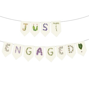 May include: A banner that reads "JUST ENGAGED" with each letter formed by flowers and greenery. The banner is made of off-white pennants strung on a thin silver wire, perfect for a wedding or engagement celebration.