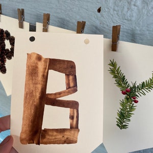 K&ouml;nnte beinhalten: Ein Banner mit dem Buchstaben "B" aus braunem Holz, zusammen mit anderen Bannern mit Naturmotiven. Das Banner wird mit einer Holzklammer vor einem hellblauen Holzgrund gehalten.