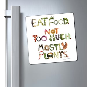 Op de afbeelding: Witte koelkastmagneet met de tekst "Eet voedsel. Niet te veel. Meestal planten." geschreven in kleurrijke letters gemaakt van verschillende fruit en groenten. De quote wordt toegeschreven aan Michael Pollan.