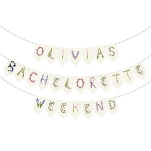 Custom floral paper banner spelling "OLIVIA'S BACHELORETTE WEEKEND" in delicate pink, sage green, and cream botanical letters. Personalized party decoration featuring pressed flower and leaf designs, perfect for bachelorette parties, bridal celebrations, and girls' weekend getaways. Elegant nature-inspired garland.