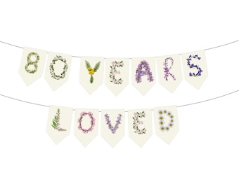 Puede incluir: Una guirnalda de banderines crema con las palabras "80 YEARS LOVED". Las letras y los números están formados con varias flores y vegetación, creando un diseño floral. Los banderines están ensartados en un alambre fino.