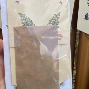 May include: A clear plastic-wrapped stationery set featuring a cream-colored banner with green sprig designs and a small brown envelope. The set includes a white card with a daisy illustration. The stationery is held by a hand.