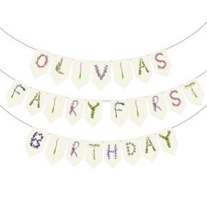 May include: A banner with three rows of cream-colored pennants strung on a thin wire. The top banner reads "OLIVIAS" in purple and green floral letters. The middle banner says "FAIRY FIRST" and the bottom banner says "BIRTHDAY".