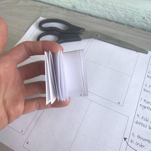 May include: A white paper booklet folded into a small book with a few pages. The booklet is being held by a hand. The image is on a white surface with black lines.